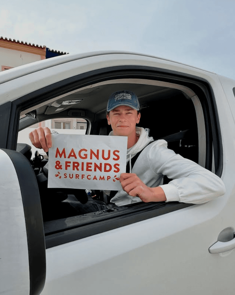 Transferchaufför håller skylt med texten "Magnus & friends surfcamp".